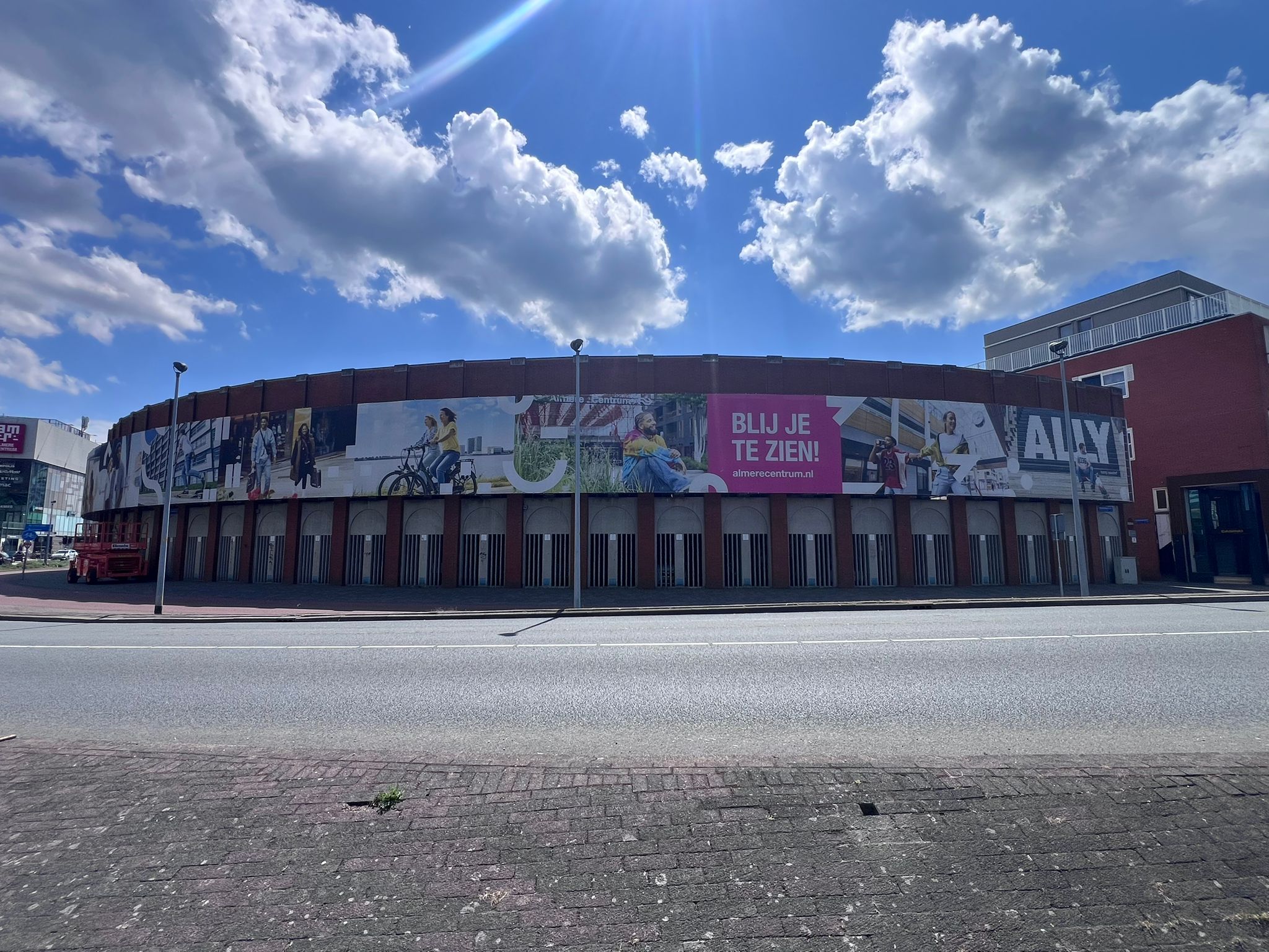 Curved brick building with large photo banner on upper front wall displaying diverse people in various activities, including cycling and sitting, with the text 'BLIJ JE TE ZIEN!' and the website almerecentrum.nl, under a blue sky with scattered clouds.