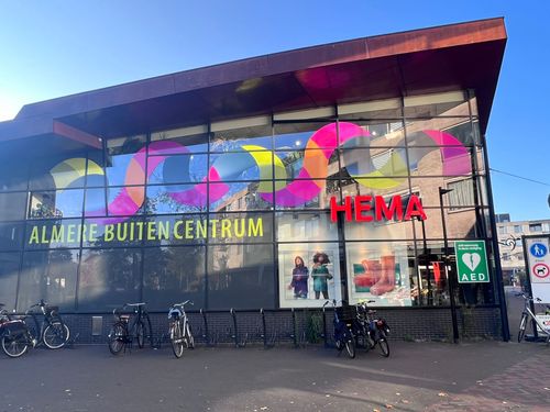 Facade of a modern building with large glass windows displaying colorful wavy stripes, the text 'ALMERE BUITEN CENTRUM' in yellow, and a red neon sign reading 'HEMA.' Several bicycles are parked in front on the pavement under a clear blue sky.