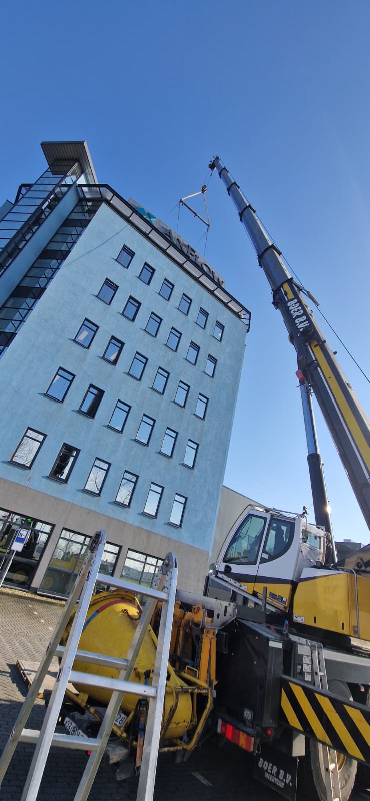Crane lifting a large sign onto the roof of a blue multi-story building under clear sky, with the crane operator cabin and equipment visible in foreground.