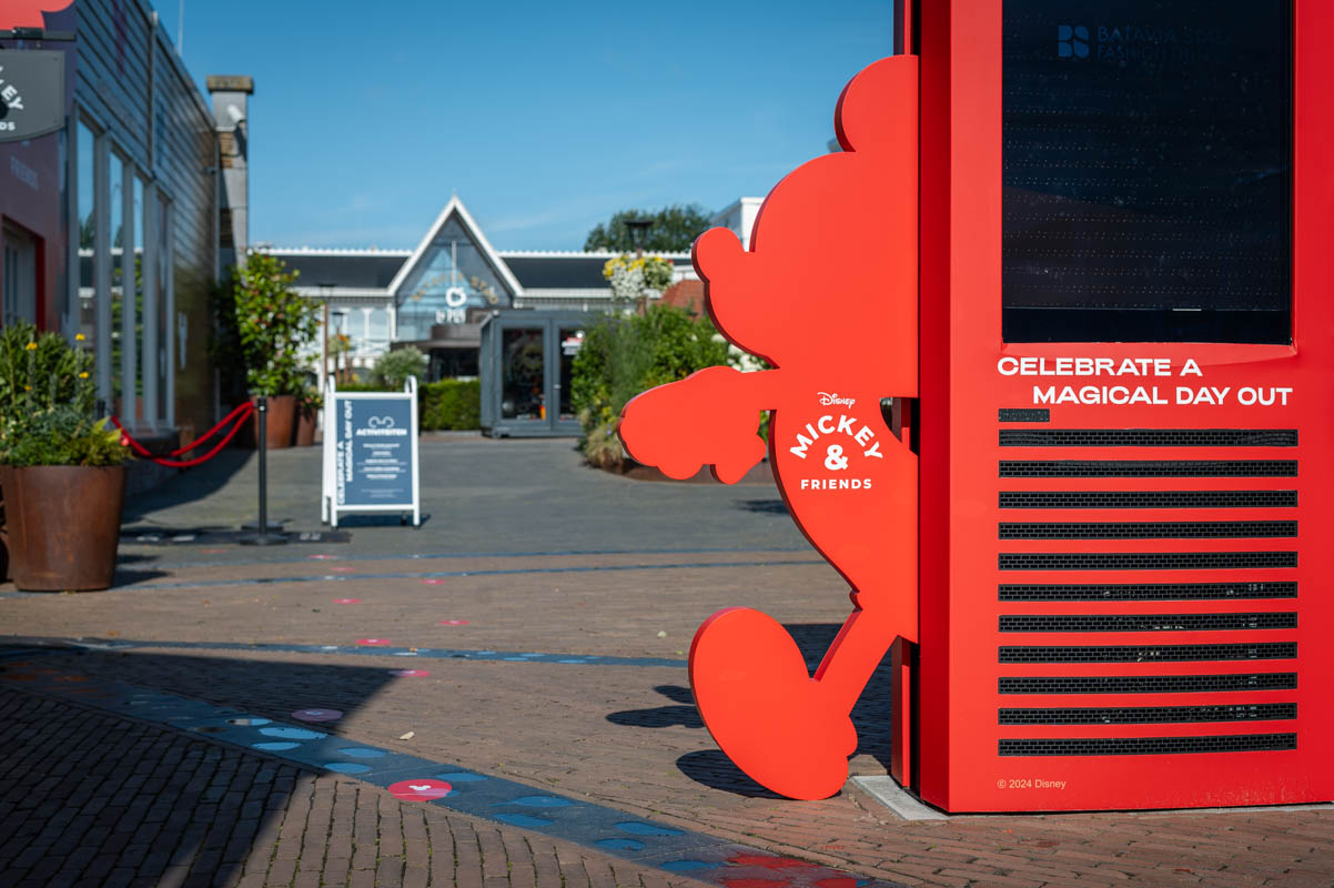 Outdoor shopping area entrance with large red display featuring Disney's Mickey & Friends silhouette and text 'Celebrate a Magical Day Out', on a sunny day.