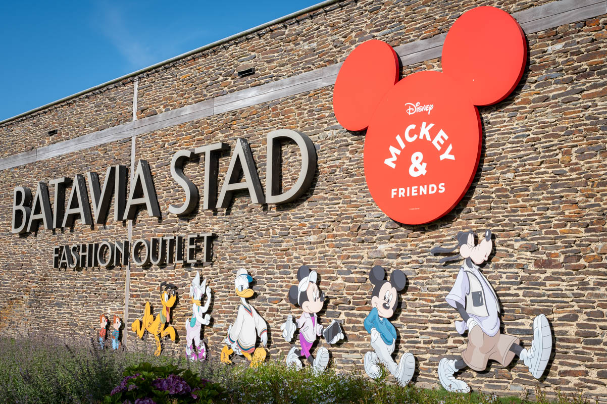 Exterior brick wall of Batavia Stad Fashion Outlet with large silver letters and a red Mickey Mouse ears sign reading 'Disney Mickey & Friends,' accompanied by colorful cutouts of Disney characters including Chip and Dale, Pluto, Daisy, Donald, Minnie, Mickey, and Goofy walking in a row.