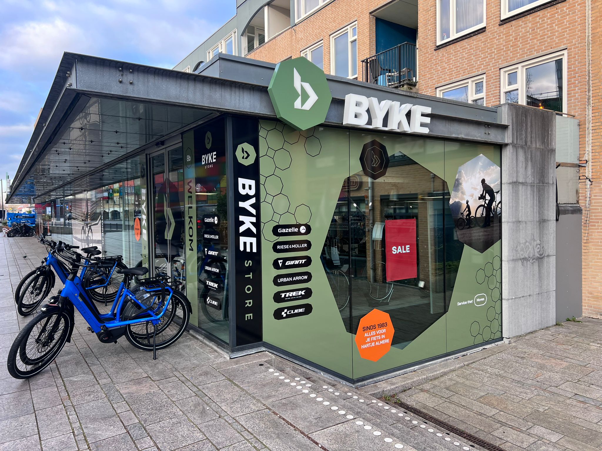 Corner storefront of BYKE Store with large glass windows and green panels displaying bicycle brand logos including Gazelle, Riese & Müller, Giant, Urban Arrow, Trek, and Cube. Blue bicycles are parked on the sidewalk outside. A red sign inside the store window reads SALE, and an orange hexagonal sign mentions service since 1983 in Dutch.