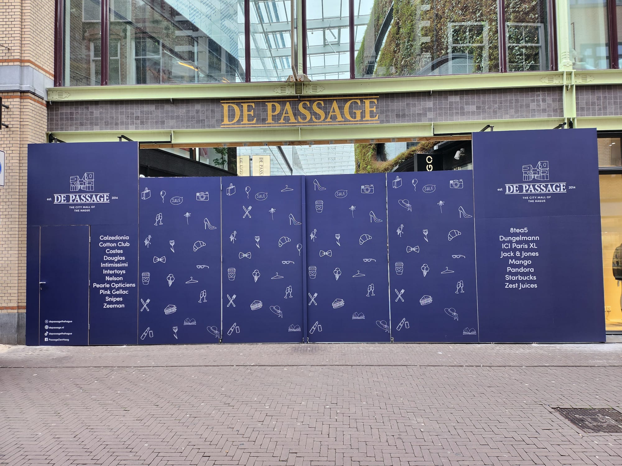 Blue construction hoarding panels in front of De Passage mall entrance with the mall's name in gold above and a list of store names printed on the hoarding under the De Passage logo, featuring icons of shopping, food, fashion, and sales scattered across the panels.