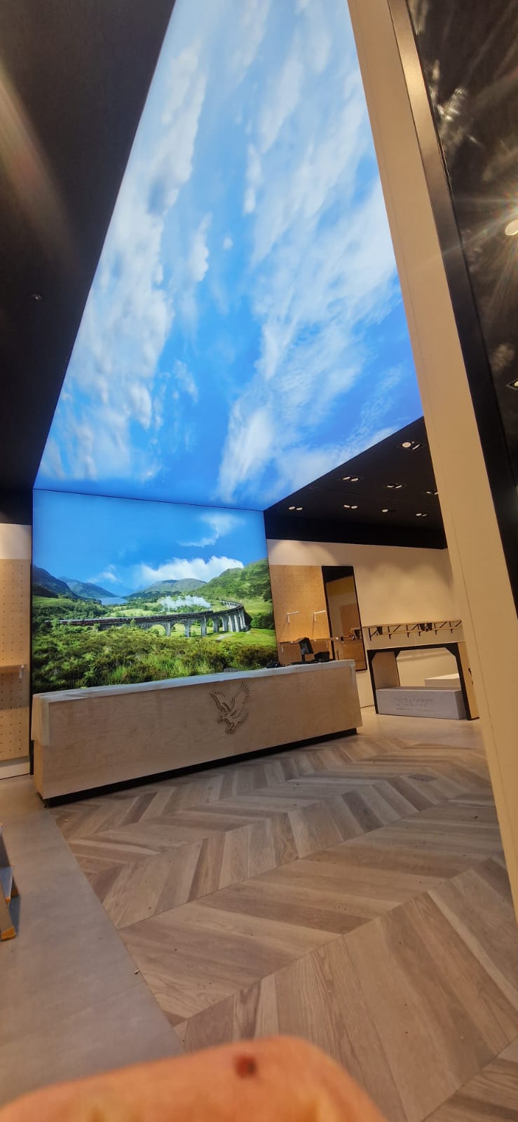 Interior of a modern retail space with light wood herringbone flooring and a light wood counter featuring a lion emblem, with a large digital sky and landscape display wrapping around the ceiling and wall showing a scenic view of a train crossing a stone viaduct in green hills under a blue sky with scattered clouds.