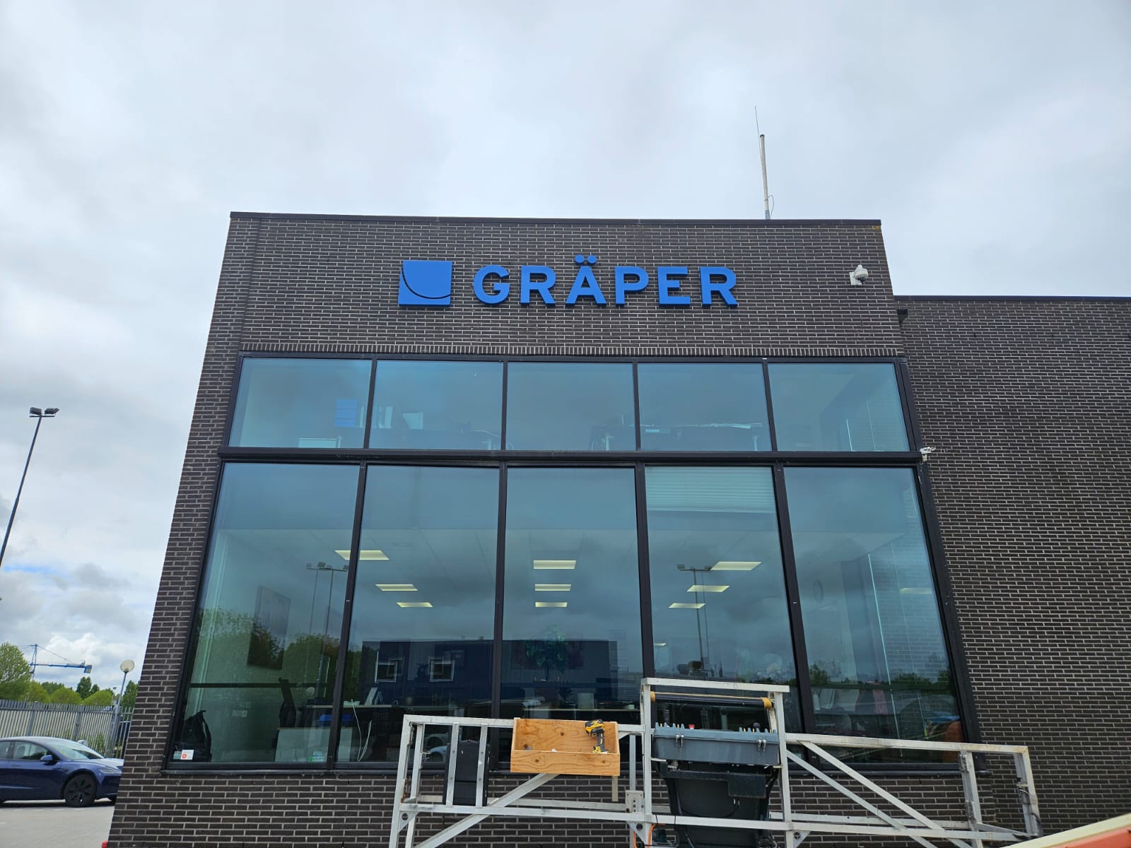 Exterior of a dark brick building with large glass windows and a blue sign reading GRÄPER above the windows under a cloudy sky.