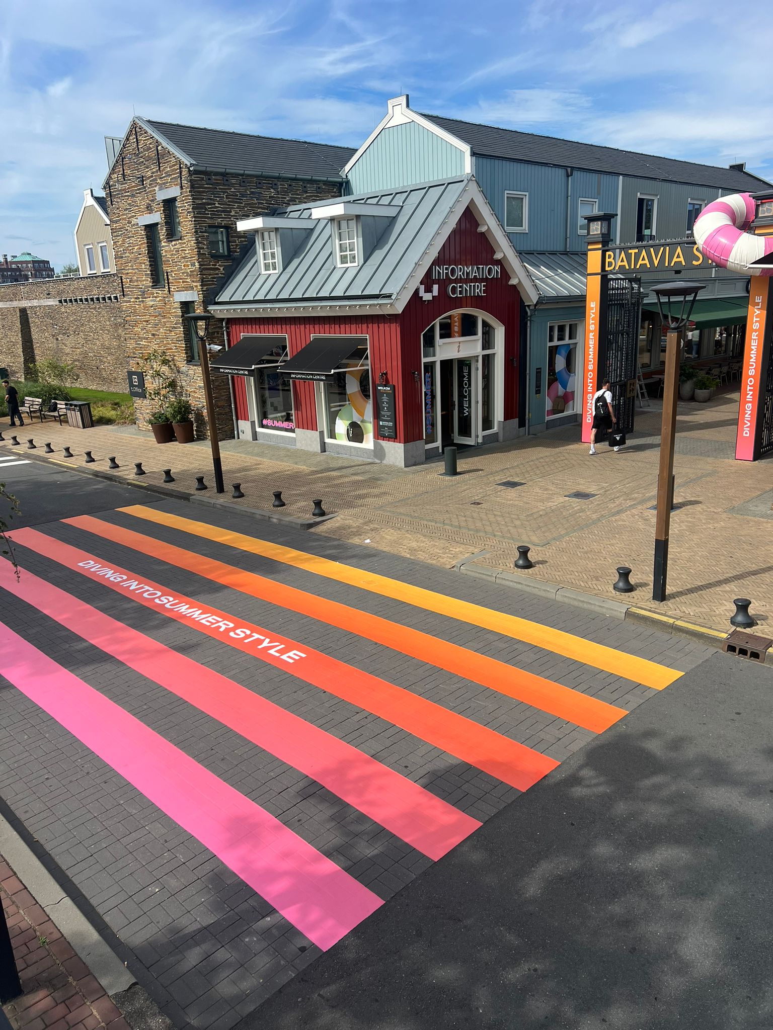 Street view of a red information centre with a gray roof next to a modern building; the street has colorful crosswalk stripes in pink, red, orange, and yellow with the text 'DIVING INTO SUMMER STYLE'.