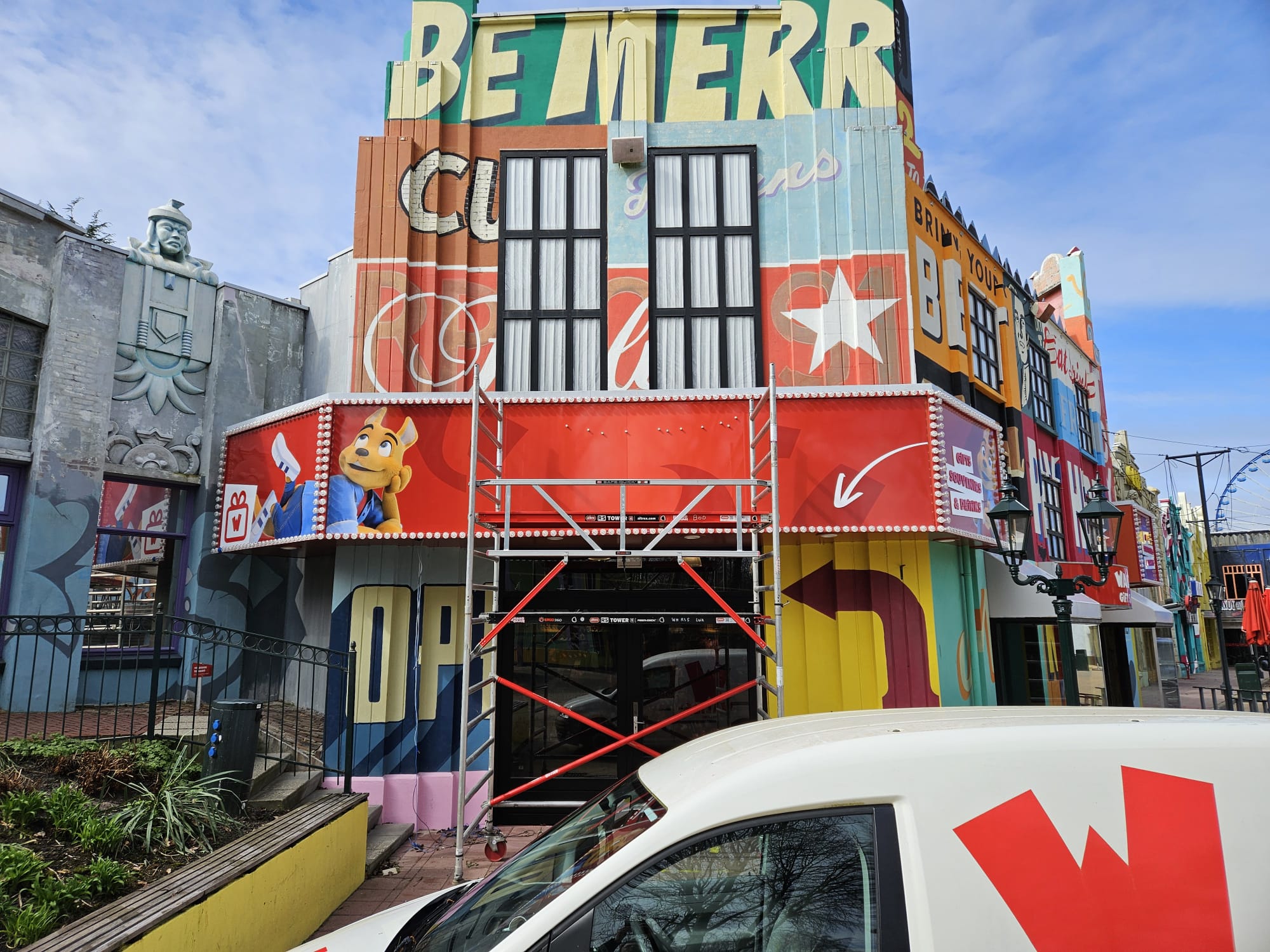 Exterior of a colorful building under scaffolding with large letters saying 'BE MERRY' at the top, a cartoon character on a red panel, and a white delivery van with red logo parked in front.