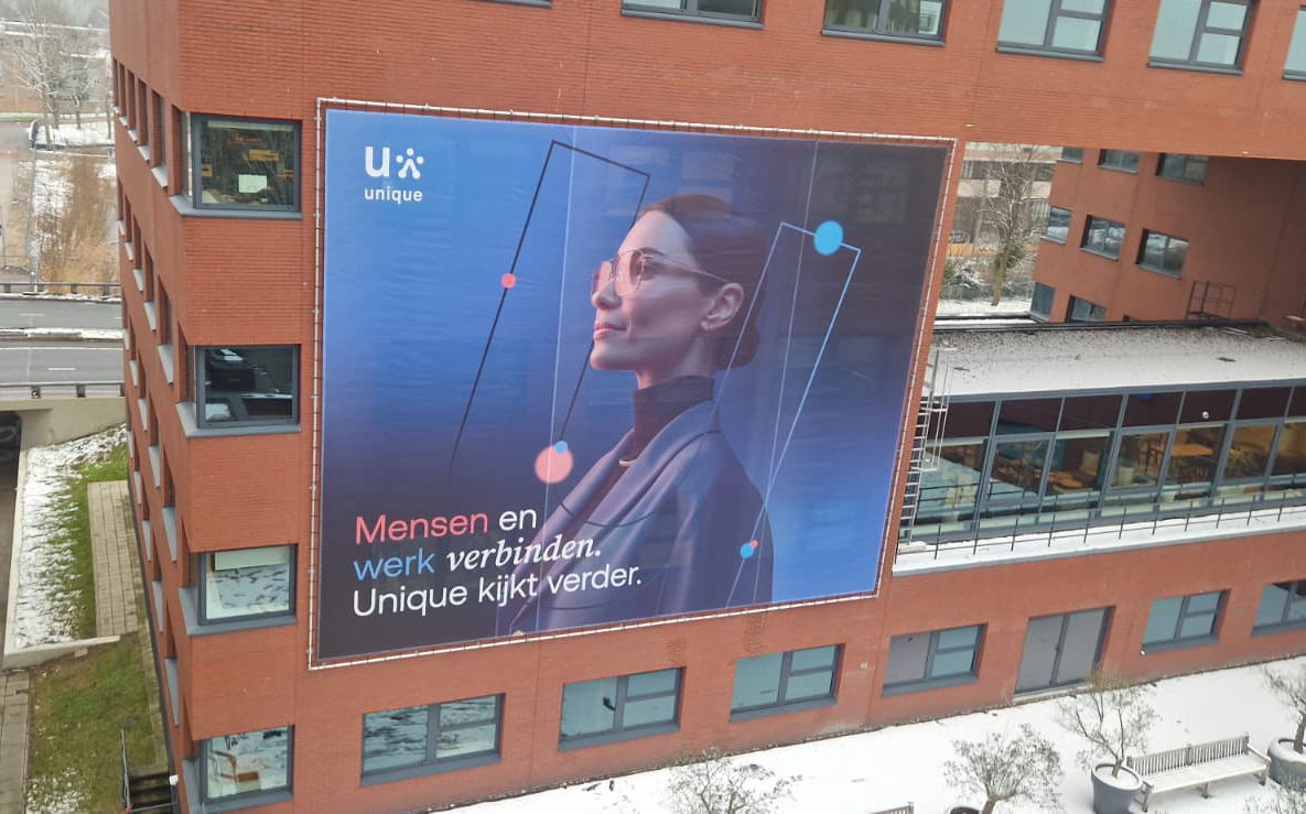 Large outdoor banner on a brick building showing a woman in profile wearing glasses and a blazer, with abstract geometric shapes around her and Dutch text that says: 'Mensen en werk verbinden. Unique kijkt verder.'