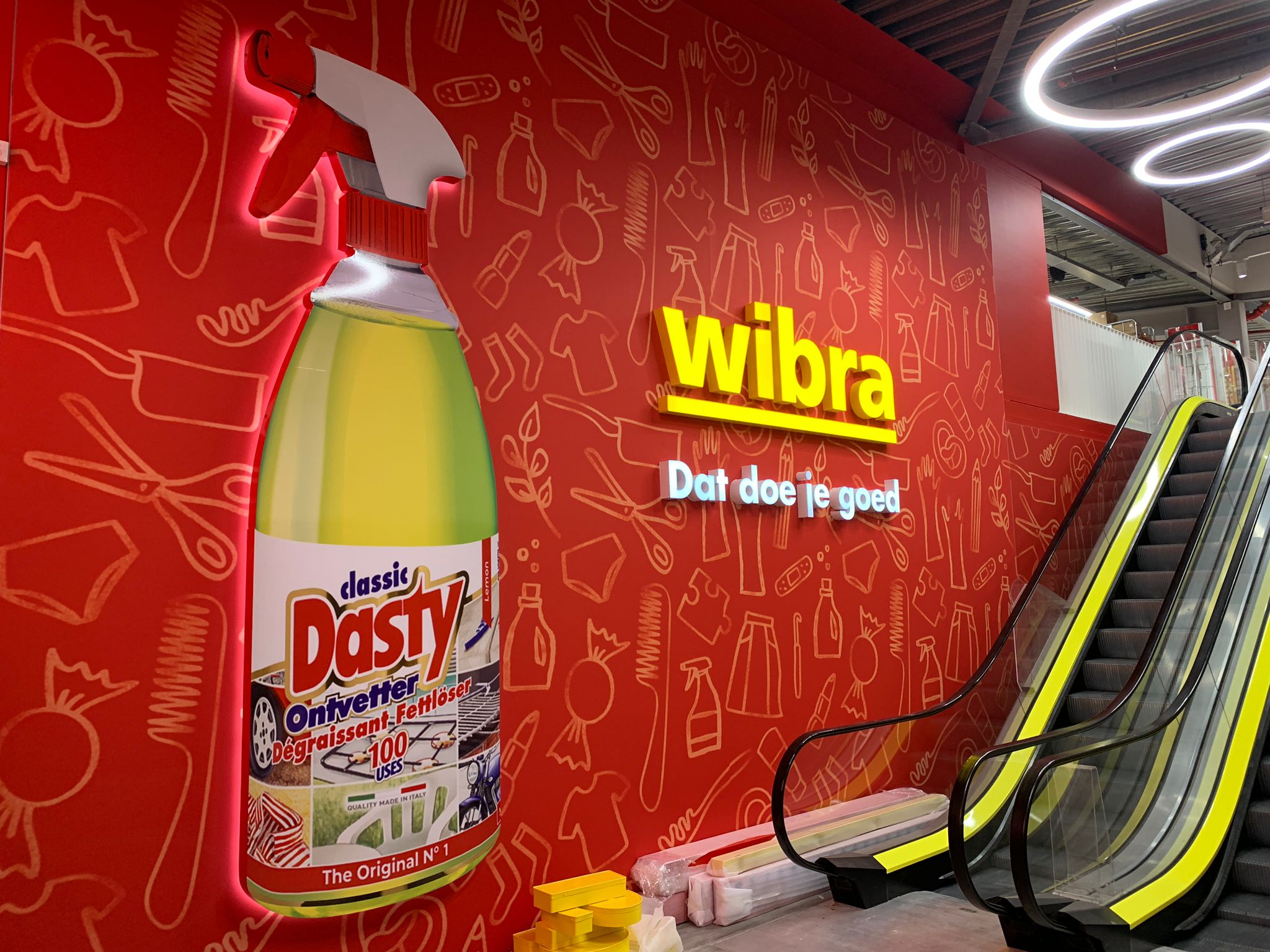 Interior wall display featuring a large, illuminated spray bottle of Dasty classic cleaning product with Dutch text, next to a bright yellow 'wibra' store sign and slogan 'Dat doe je goed' on a red background with line illustrations of clothing and household items, beside an escalator with yellow accents.