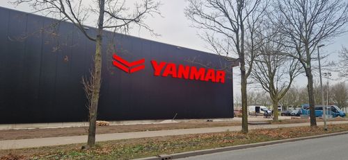 Black building exterior with large red illuminated YANMAR logo on the side; leafless trees and parked vehicles are visible nearby.