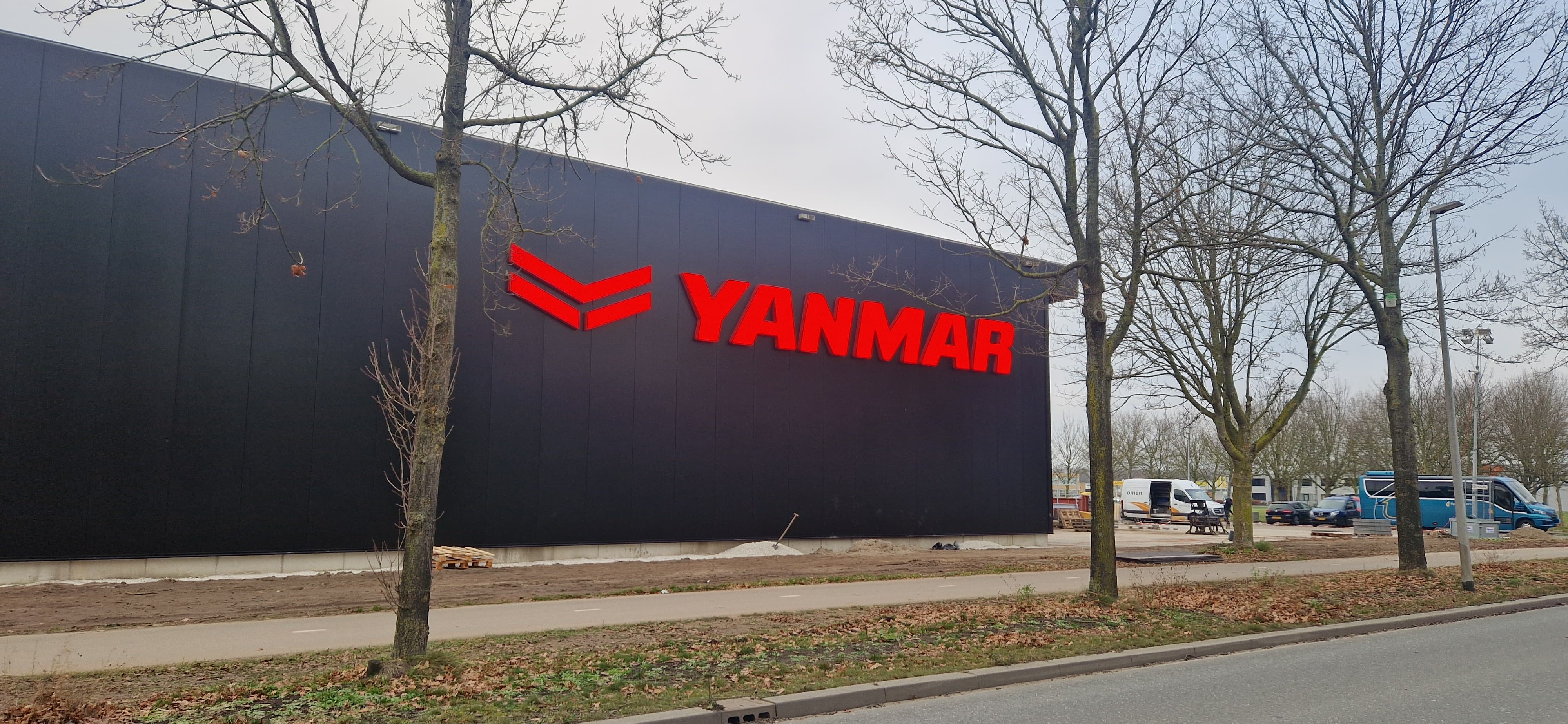 Black building exterior with large red illuminated YANMAR logo on the side; leafless trees and parked vehicles are visible nearby.