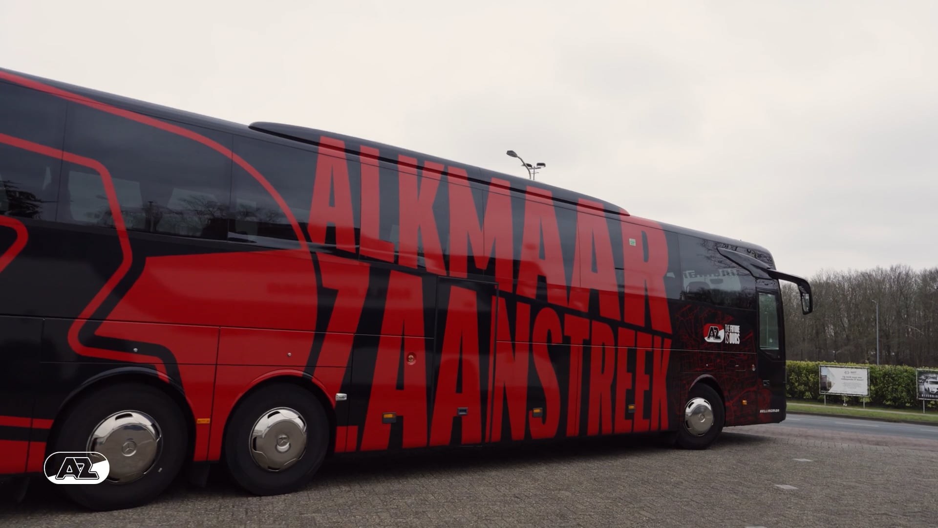 Zijaanzicht van een bus met grote rode letters 'ALKMAAR ZAANSTREEK' op een zwarte en rood gestreepte achtergrond, geparkeerd op een betegelde weg met bomen en reclameborden op afstand.