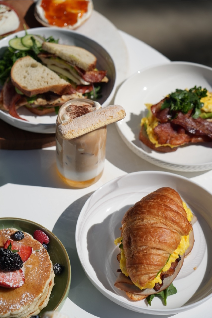 Table with breakfast items including a layered sandwich with bacon and greens, scrambled eggs with bacon on a plate, a croissant sandwich with eggs and meat, a stack of pancakes topped with berries, and an iced coffee with a biscotti.
