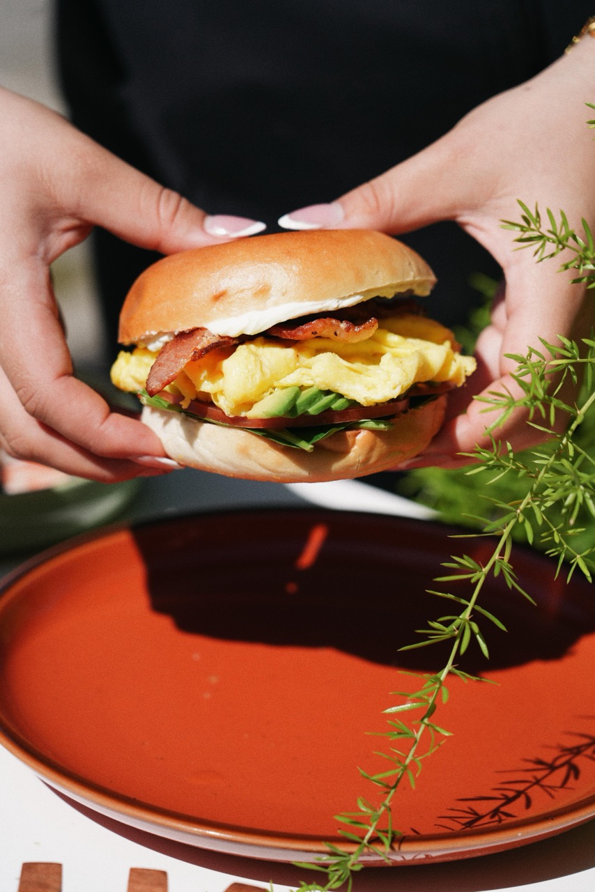 Hands holding a breakfast sandwich with scrambled eggs, bacon, avocado, tomato, and greens on a toasted bun above a red plate.
