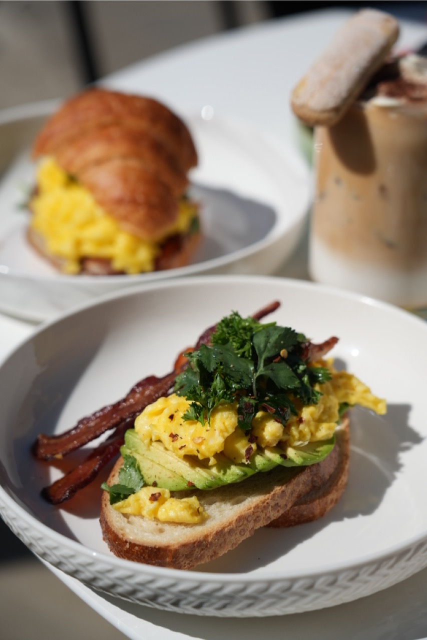 White plate with toast topped with avocado slices, scrambled eggs, fresh herbs, and two strips of crispy bacon, with a croissant sandwich and iced coffee in the blurred background.