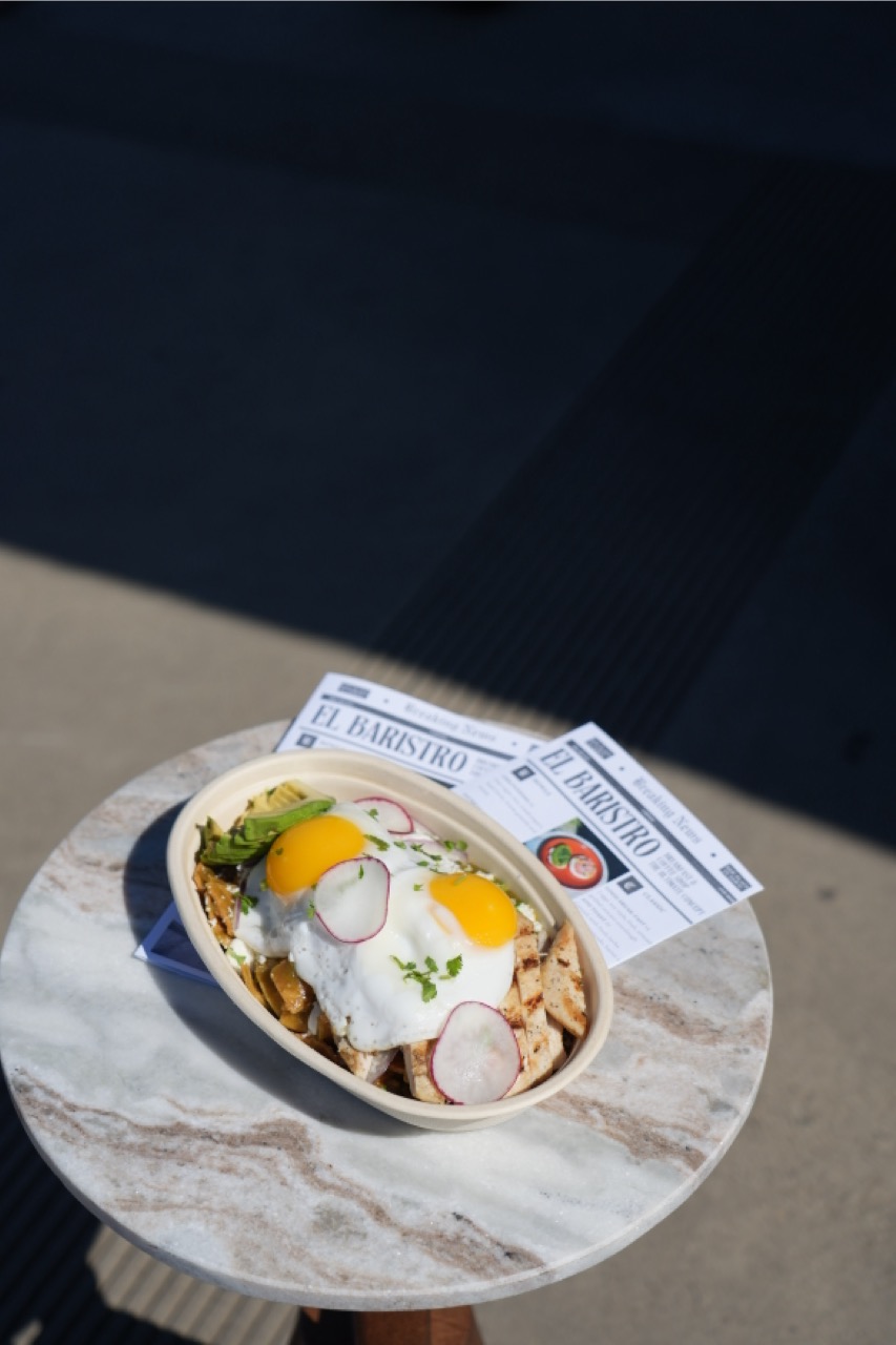 Bowl with grilled chicken strips, two sunny-side-up eggs topped with radish slices and herbs, and avocado slices on the side, placed on a round marble table with two El Baristro flyers underneath.