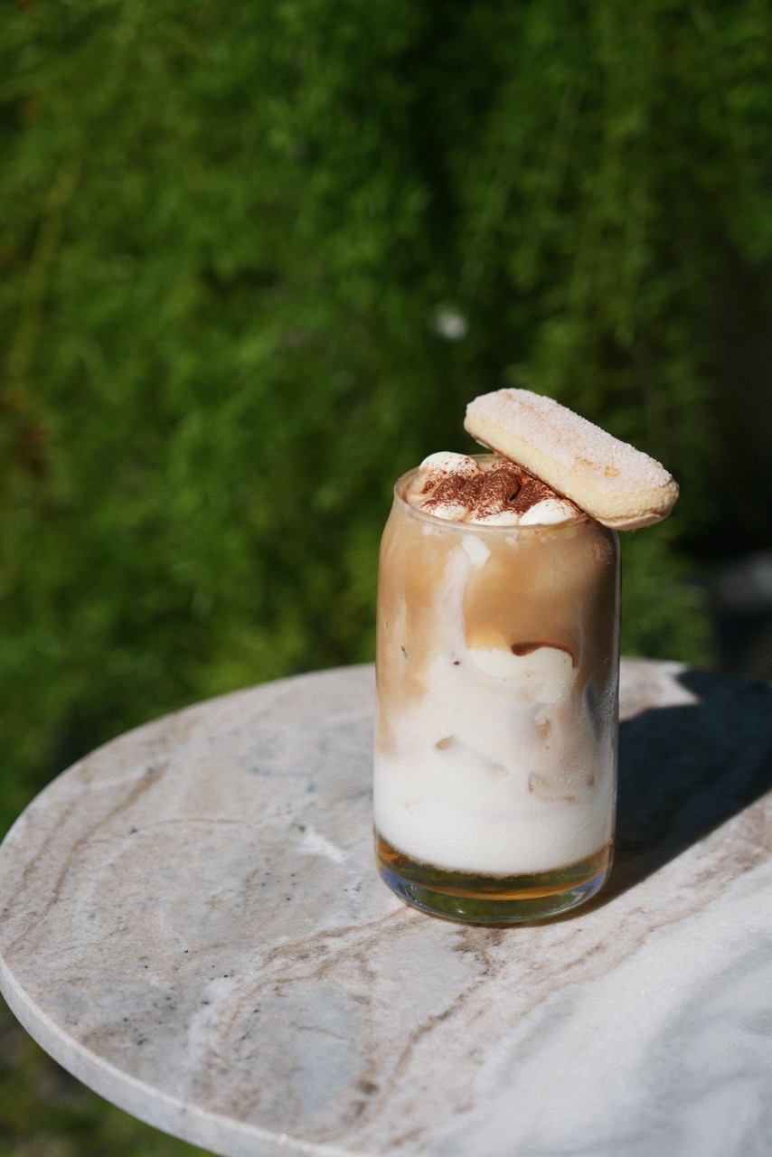 Iced coffee with a layered look of milk and coffee topped with whipped cream and cocoa powder, garnished with a ladyfinger biscuit, on a round marble table with a blurred green background.