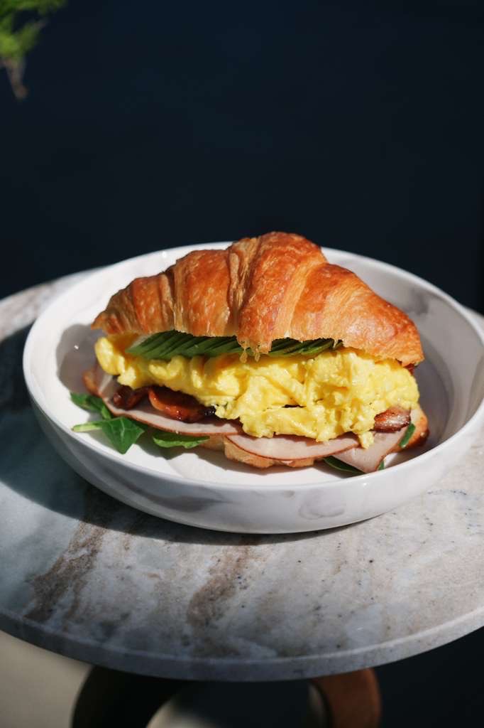 Croissant sandwich with scrambled eggs, sliced turkey, bacon, spinach, and sliced avocado on a white plate on a marble table.