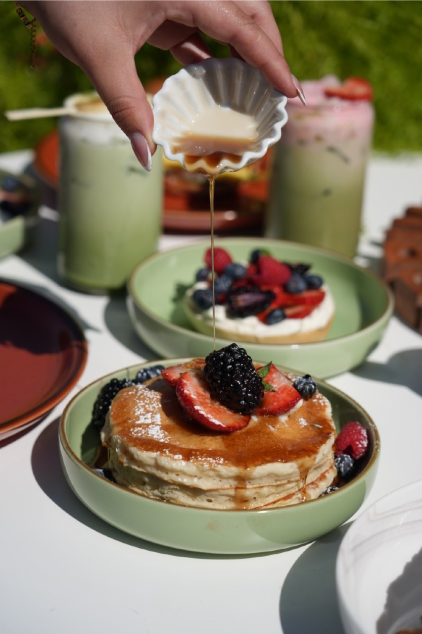 Hand pouring syrup over stack of pancakes topped with strawberries and blackberries in a green bowl, with another bowl of fruit-topped dessert and two drinks blurred in background.