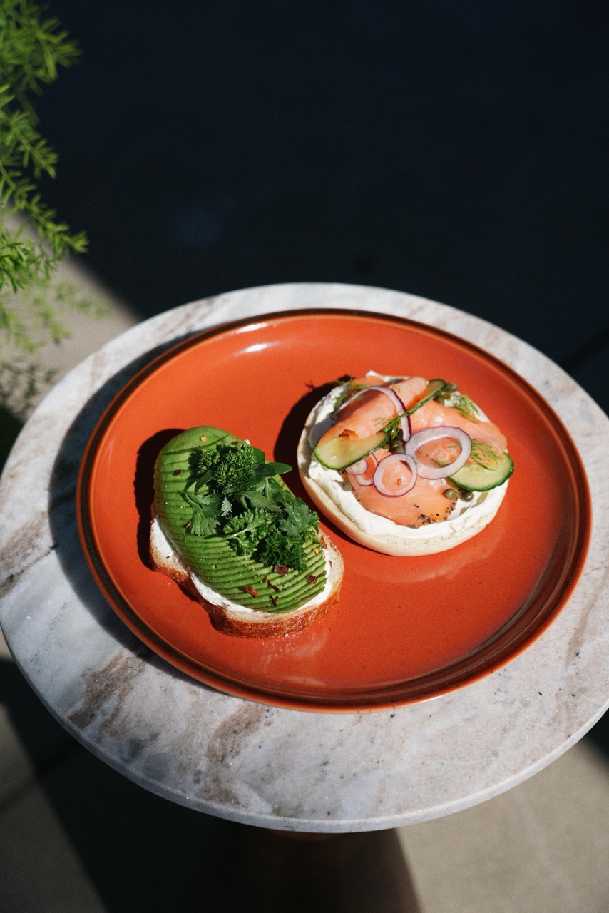 Orange plate on marble table with two open-faced sandwiches: one topped with sliced avocado and herbs, the other with smoked salmon, cucumber slices, red onion rings, and capers on cream cheese.