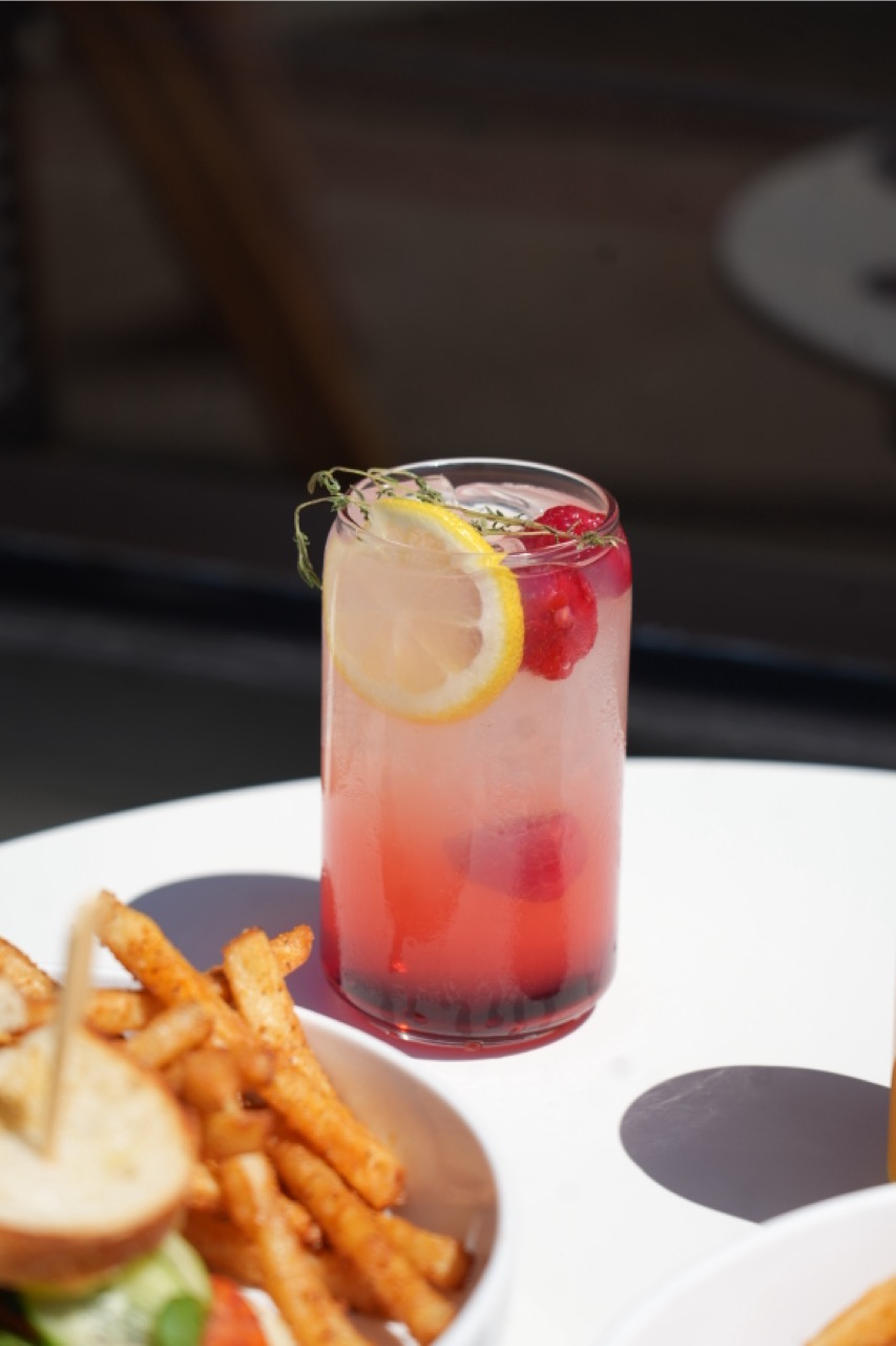A glass of pink lemonade with ice, raspberries, a lemon slice, and thyme garnish on a white table next to a plate of seasoned French fries and a sandwich.
