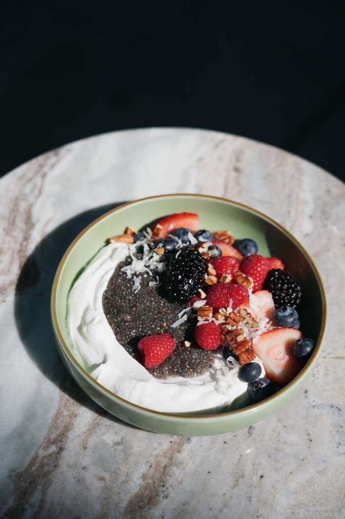 Bowl of chia seed pudding topped with whipped cream, blackberries, raspberries, blueberries, strawberry slices, shredded coconut, and chopped pecans on a marble surface.