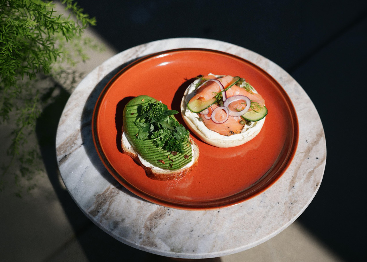 Open-faced bagel halves on an orange plate atop a marble table, one with sliced avocado and herbs, the other with smoked salmon, cream cheese, cucumber slices, red onion, and capers.