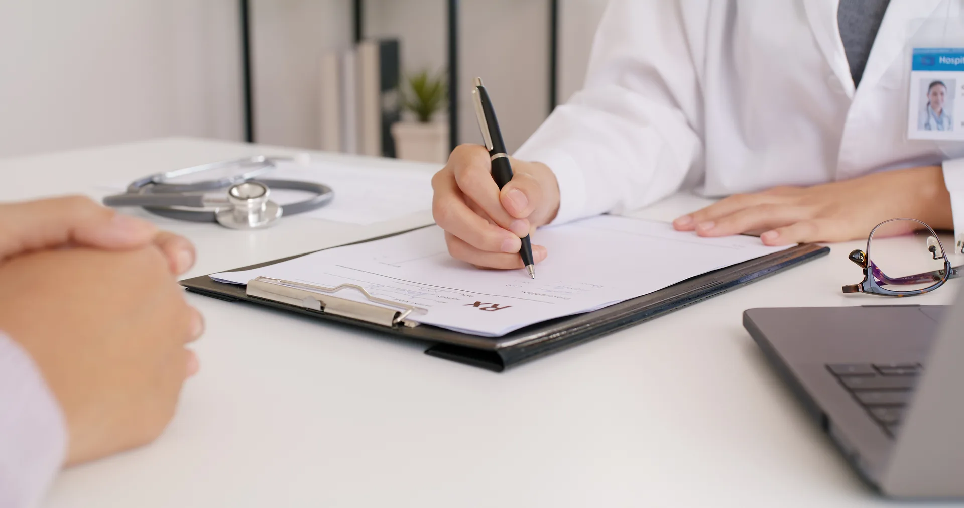 A clinician writing a prescription on a clipboard across the desk from a patient