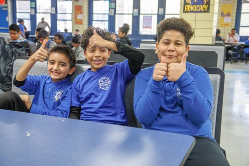 Three students in blue uniforms giving thumbs up in cafeteria