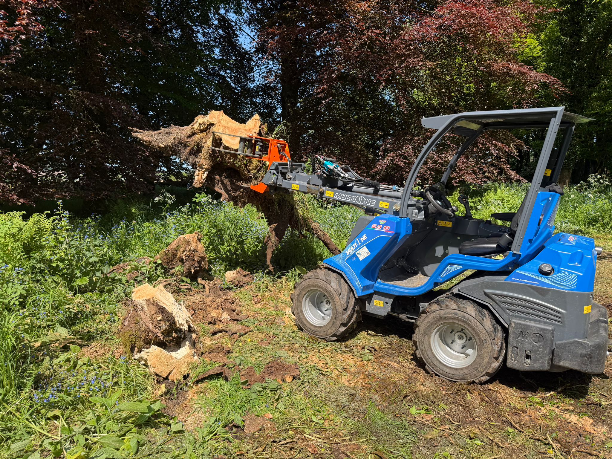 Blue MultiOne compact loader lifting a large tree stump with roots in a grassy, wooded area.