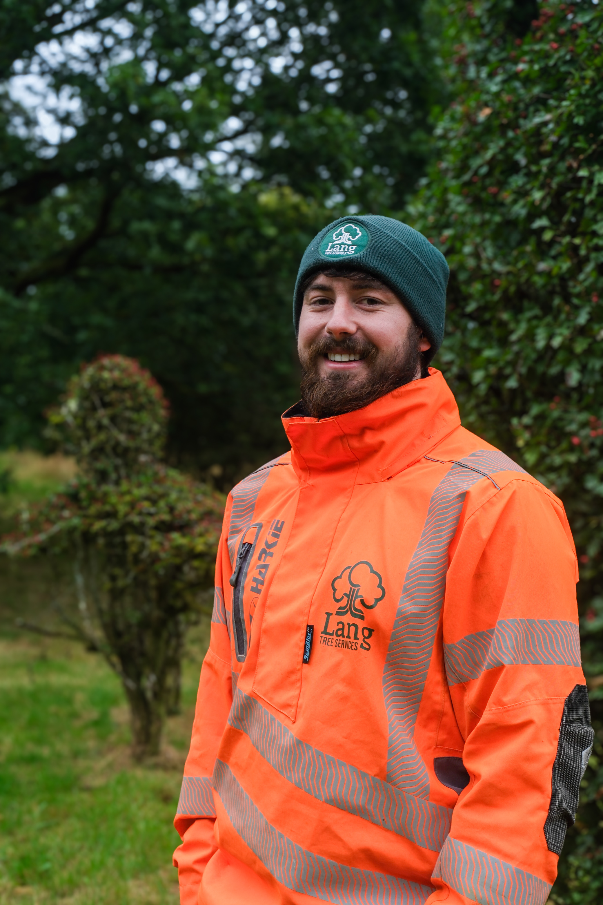 Smiling man in an orange high-visibility jacket and green beanie with Lang Tree Services logo outdoors.