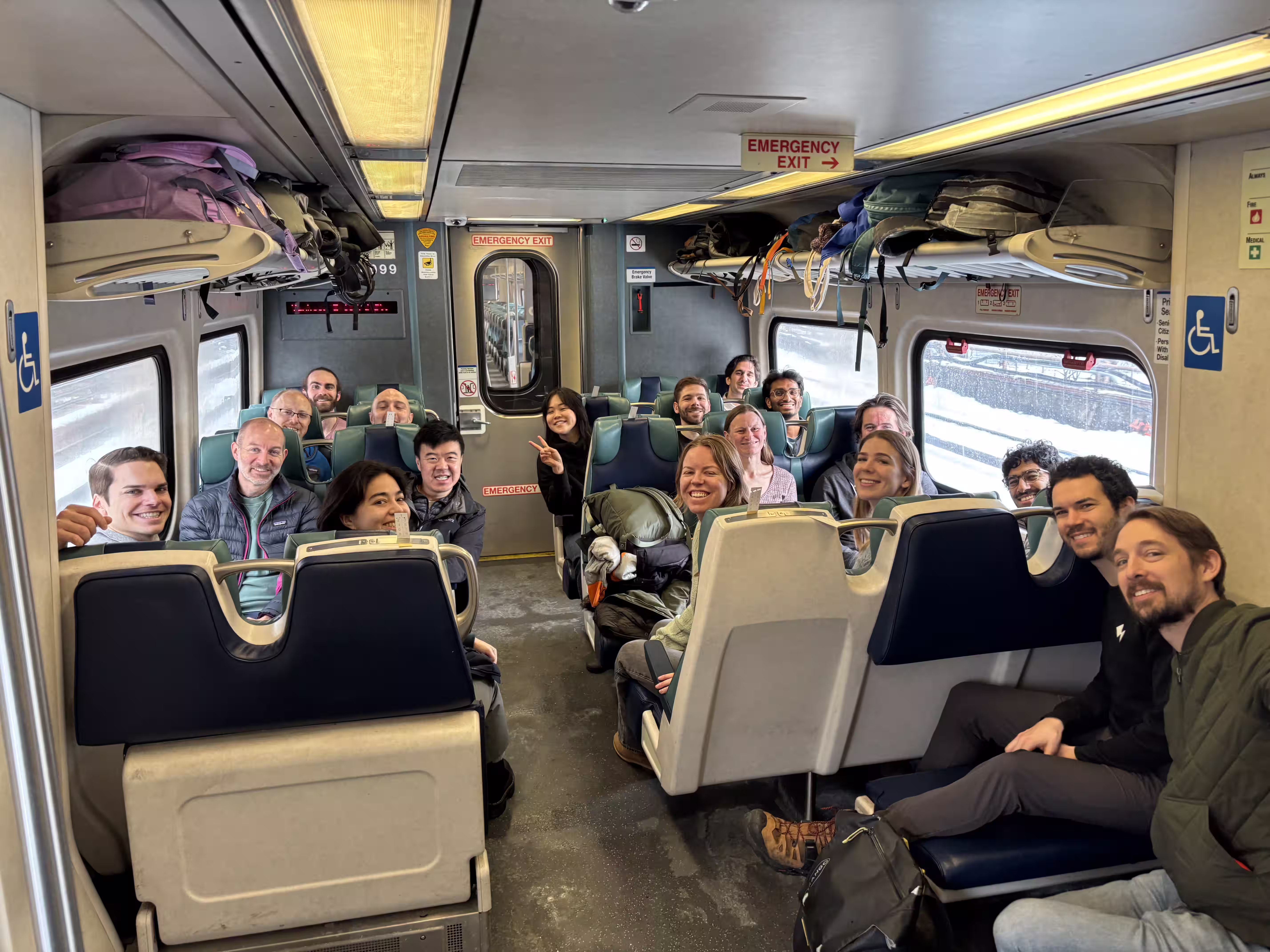 Group of smiling people seated facing each other inside a train car with luggage overhead.