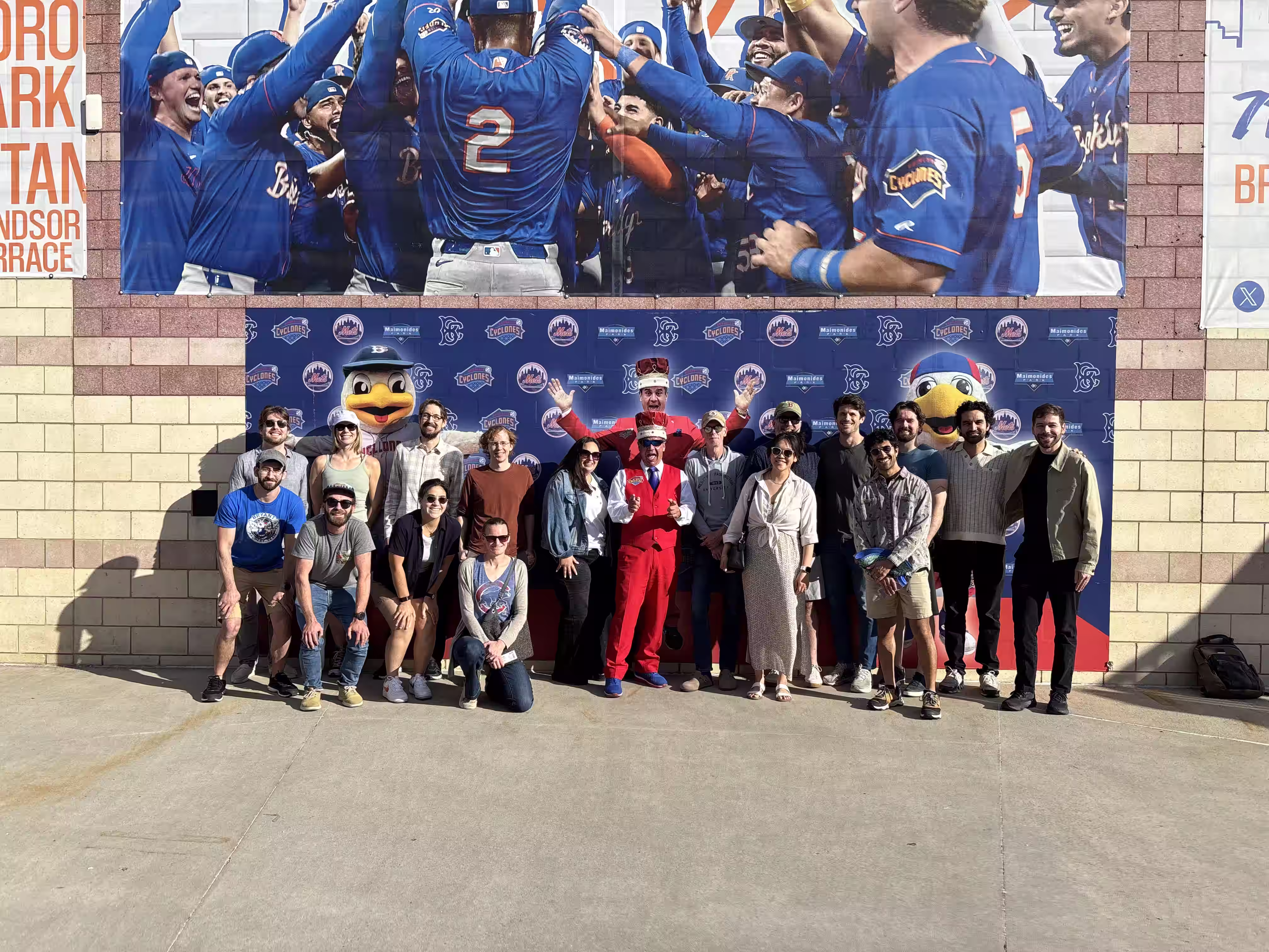 Group of people posing outdoors in front of a blue wall with sports logos and mascots.