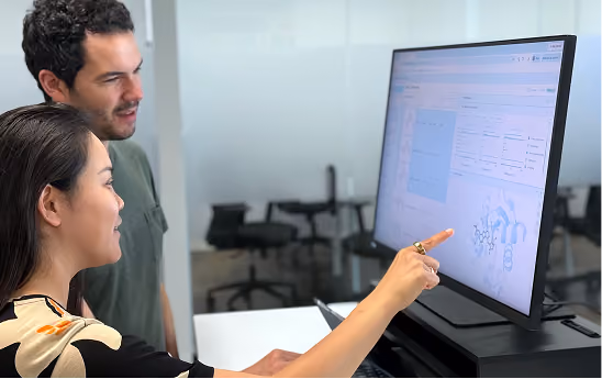 Two people, a woman in the foreground pointing at a large computer screen displaying a software interface with charts and diagrams, and a man standing next to her watching attentively in an office setting.