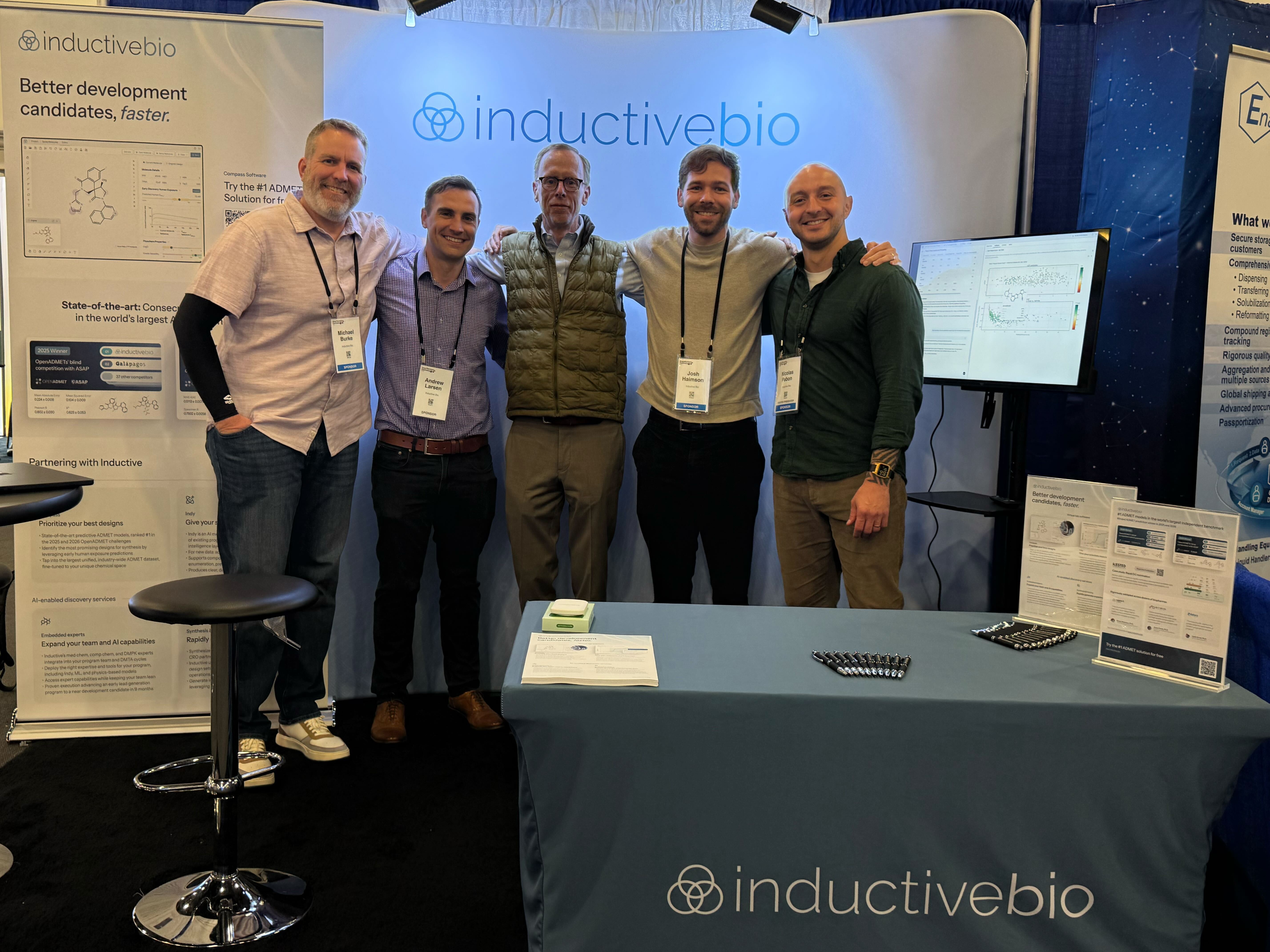 Five men standing side by side, smiling, at an InductiveBio booth with banners and a table displaying company information and pens.