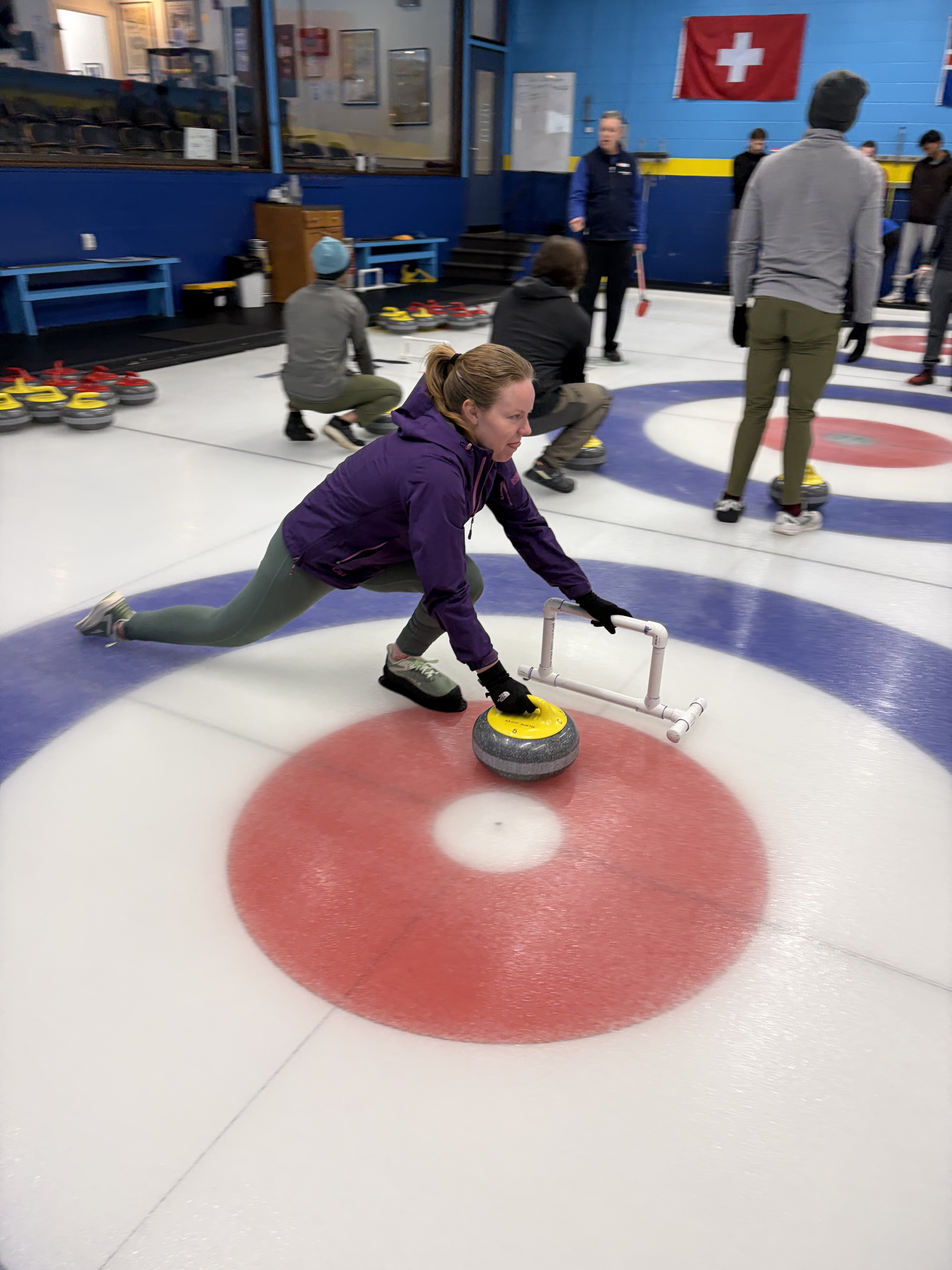 Woman in purple jacket and green leggings sliding on ice while pushing a yellow-handled curling stone in an indoor curling rink with other players and curling stones visible in the background.