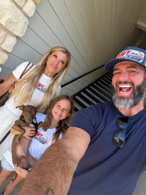 Smiling man with beard wearing a cap takes a selfie with a woman, a girl holding a small brown dog, all wearing matching white and navy shirts with a logo, standing outside near stone and wood siding with stairs behind.