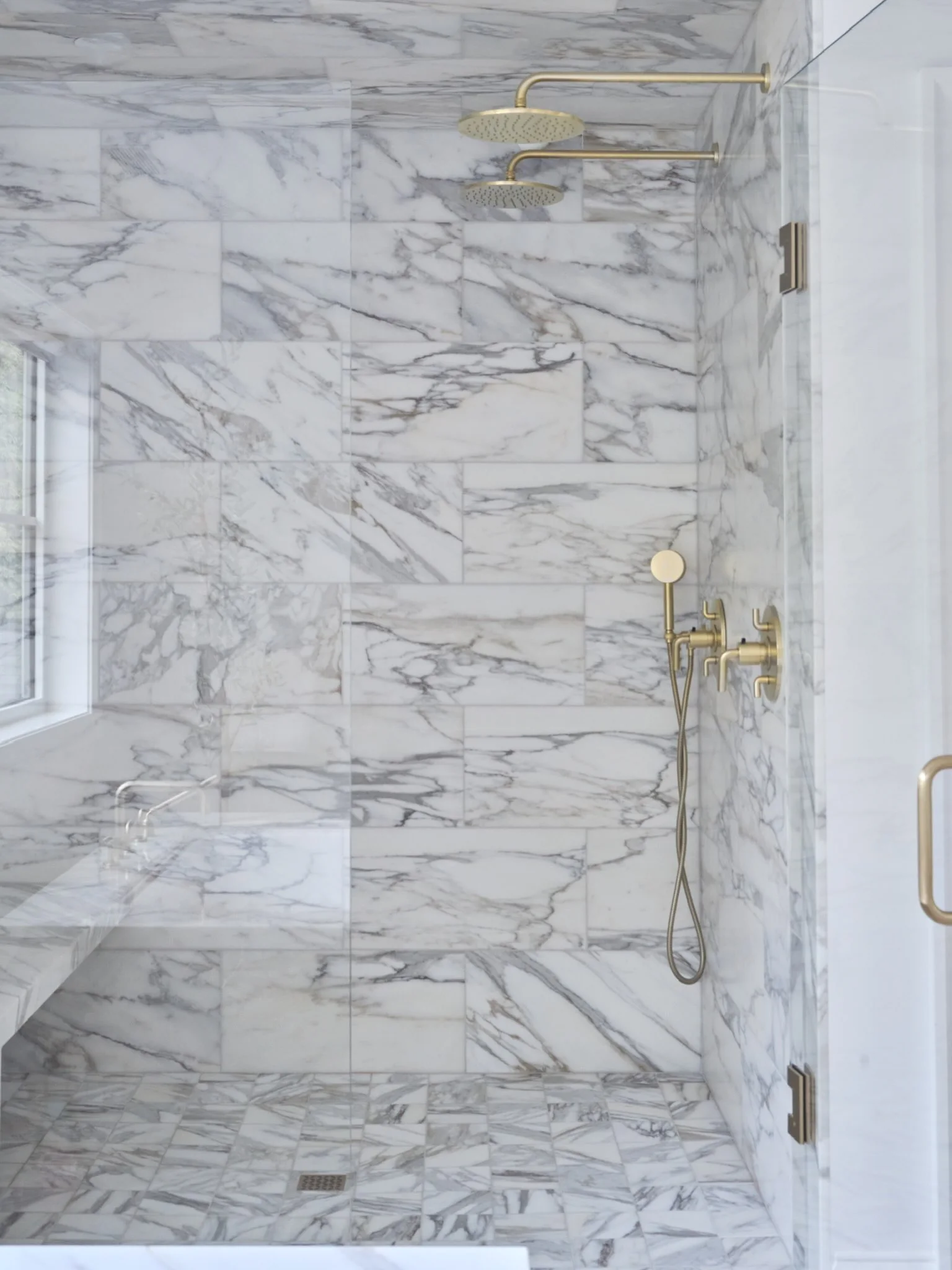 Marble shower with a stone bench, two brass rain shower heads and glass enclosure.