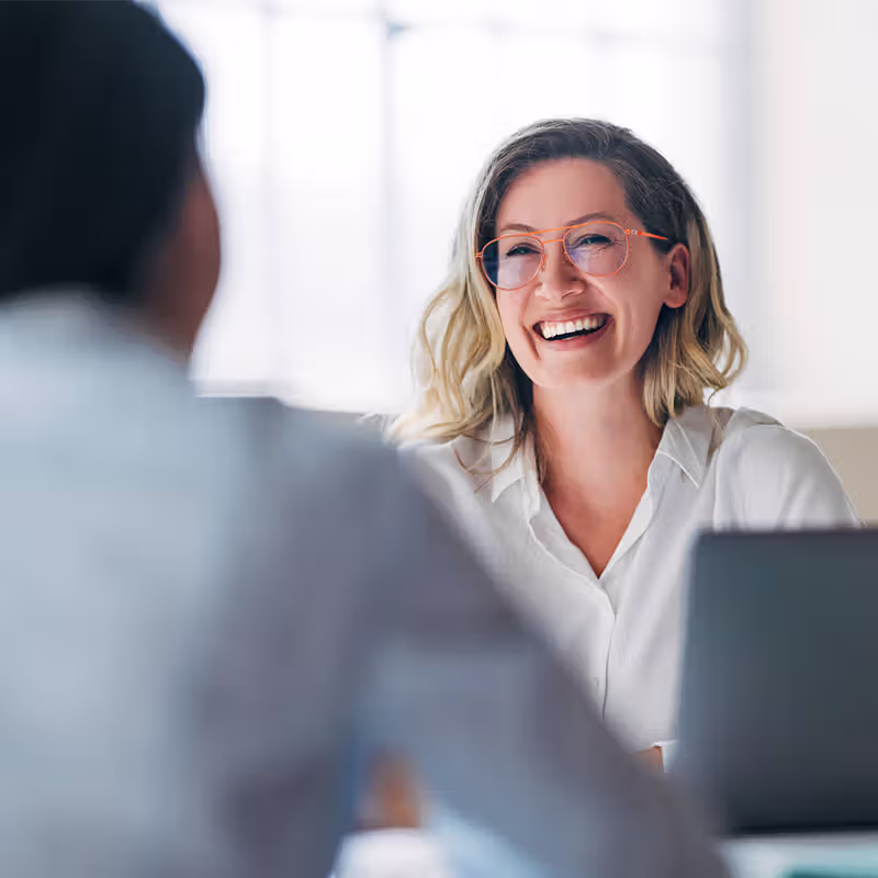 Smiling woman with glasses and blonde hair talking to a person in an office setting.
