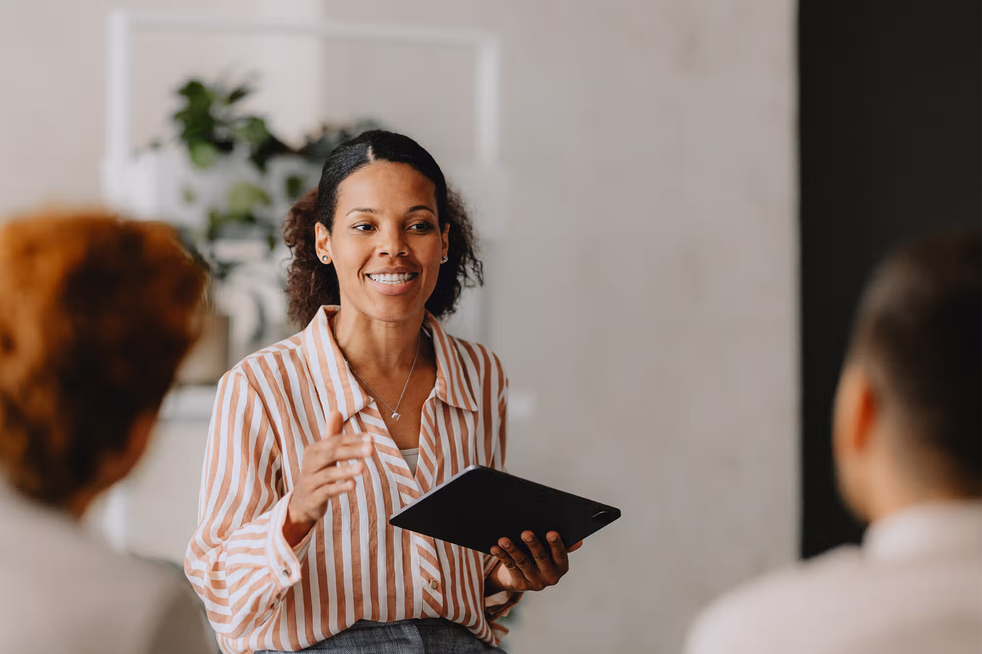 Smiling woman in a striped shirt holding a tablet and speaking to two people in a meeting.