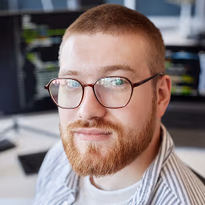 Close-up of a young man with glasses, beard, and short hair looking at the camera in a tech workspace.