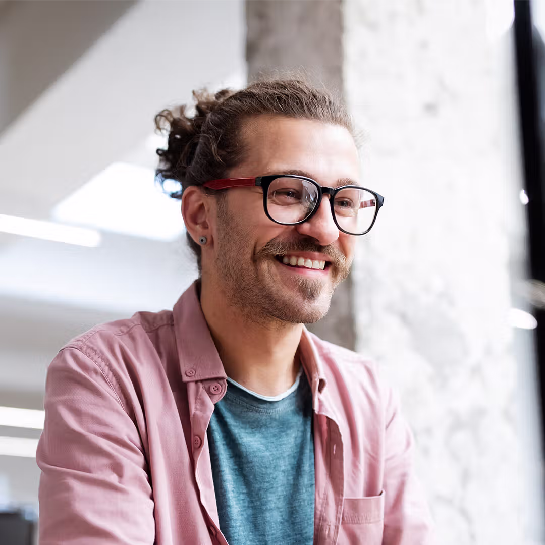 Smiling man with glasses, mustache, and tied-back hair wearing a pink shirt over a blue t-shirt indoors.