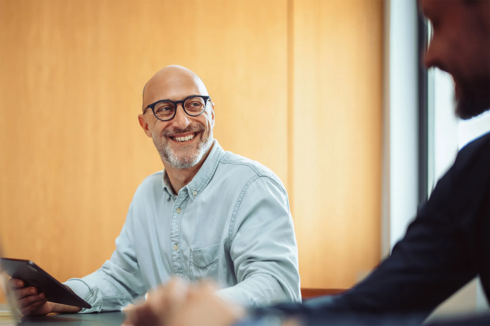 Smiling bald man with glasses holding a tablet and looking towards a person in a dark suit.