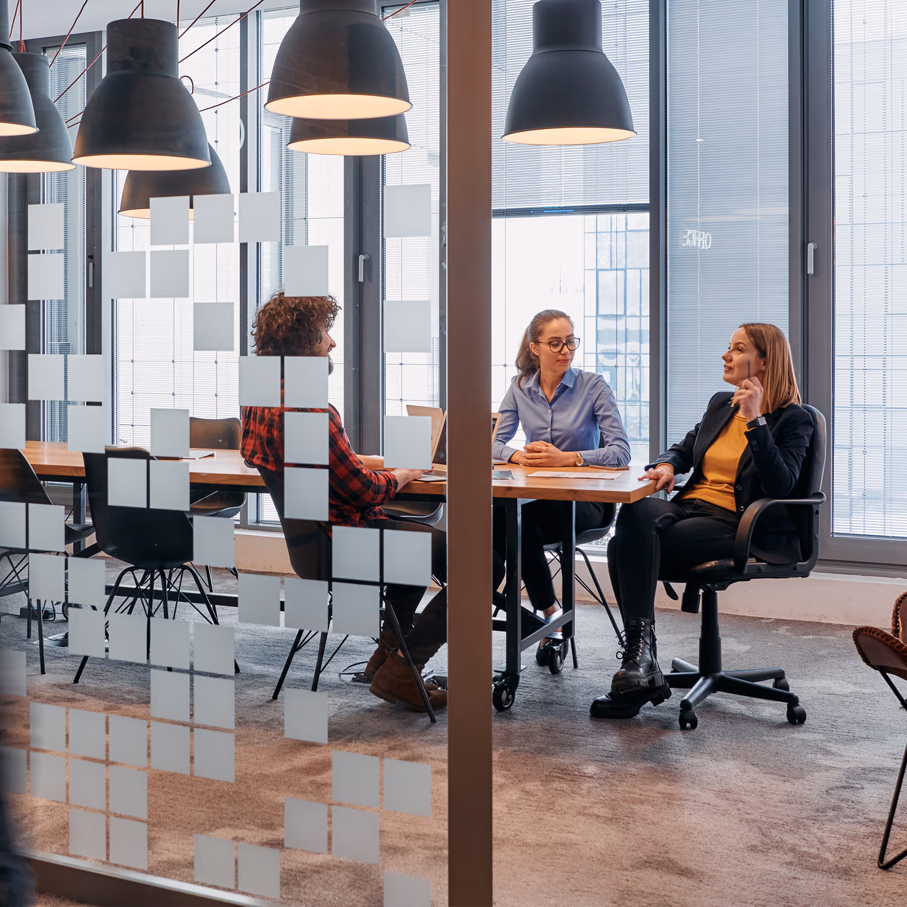 Three colleagues sitting around an office table having a discussion in a modern meeting room with large windows.
