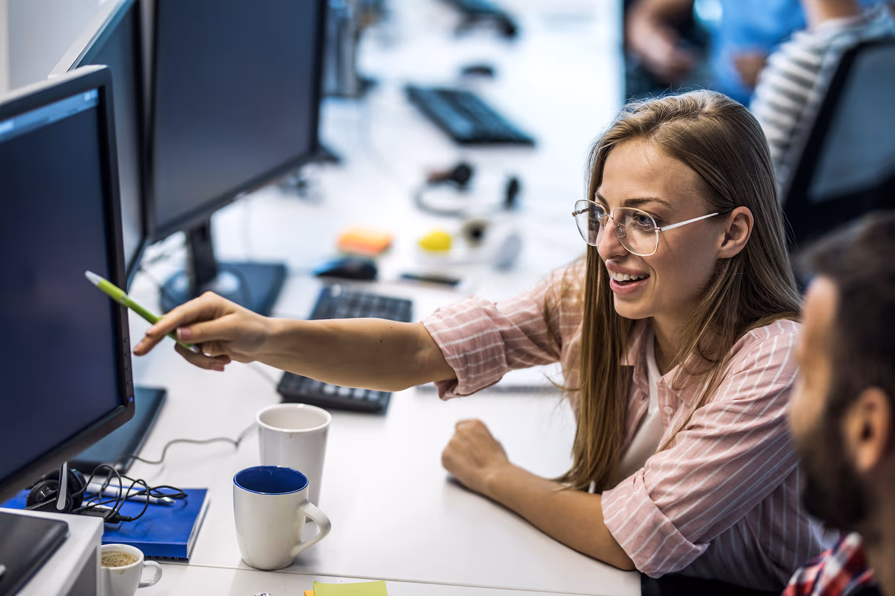 Woman in glasses pointing at a computer screen with a pen while talking to a man at a shared office desk.