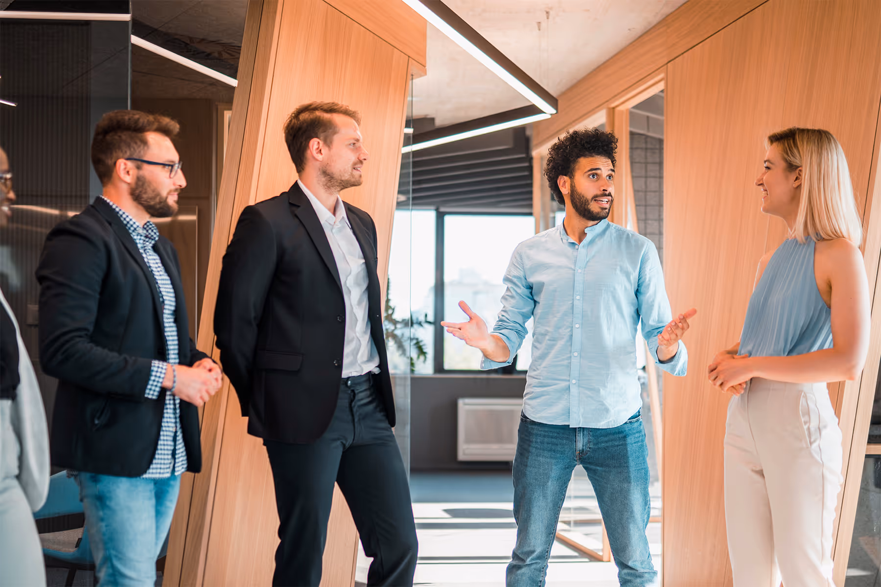 Diverse group of four professionals standing and having a discussion in a modern office hallway with wooden walls.