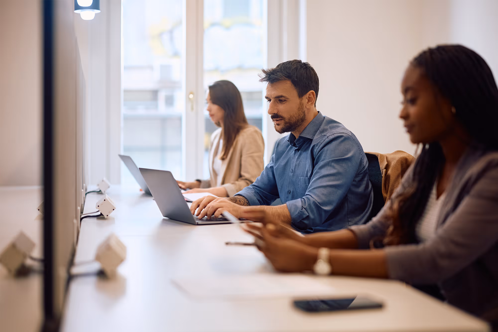 Three professionals working at laptops and a smartphone in a bright office with large windows.