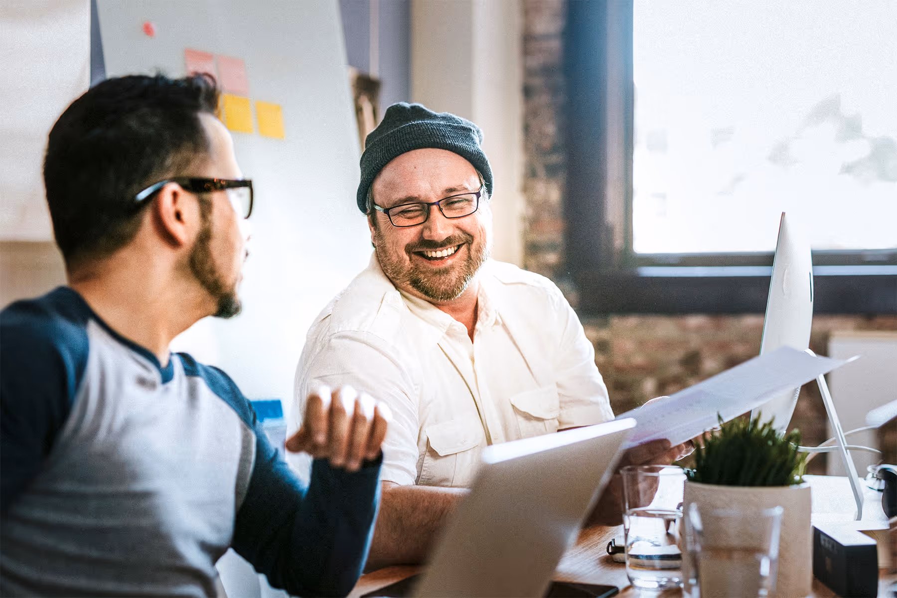 Two men in an office discussing documents, one wearing glasses and a beanie, smiling.