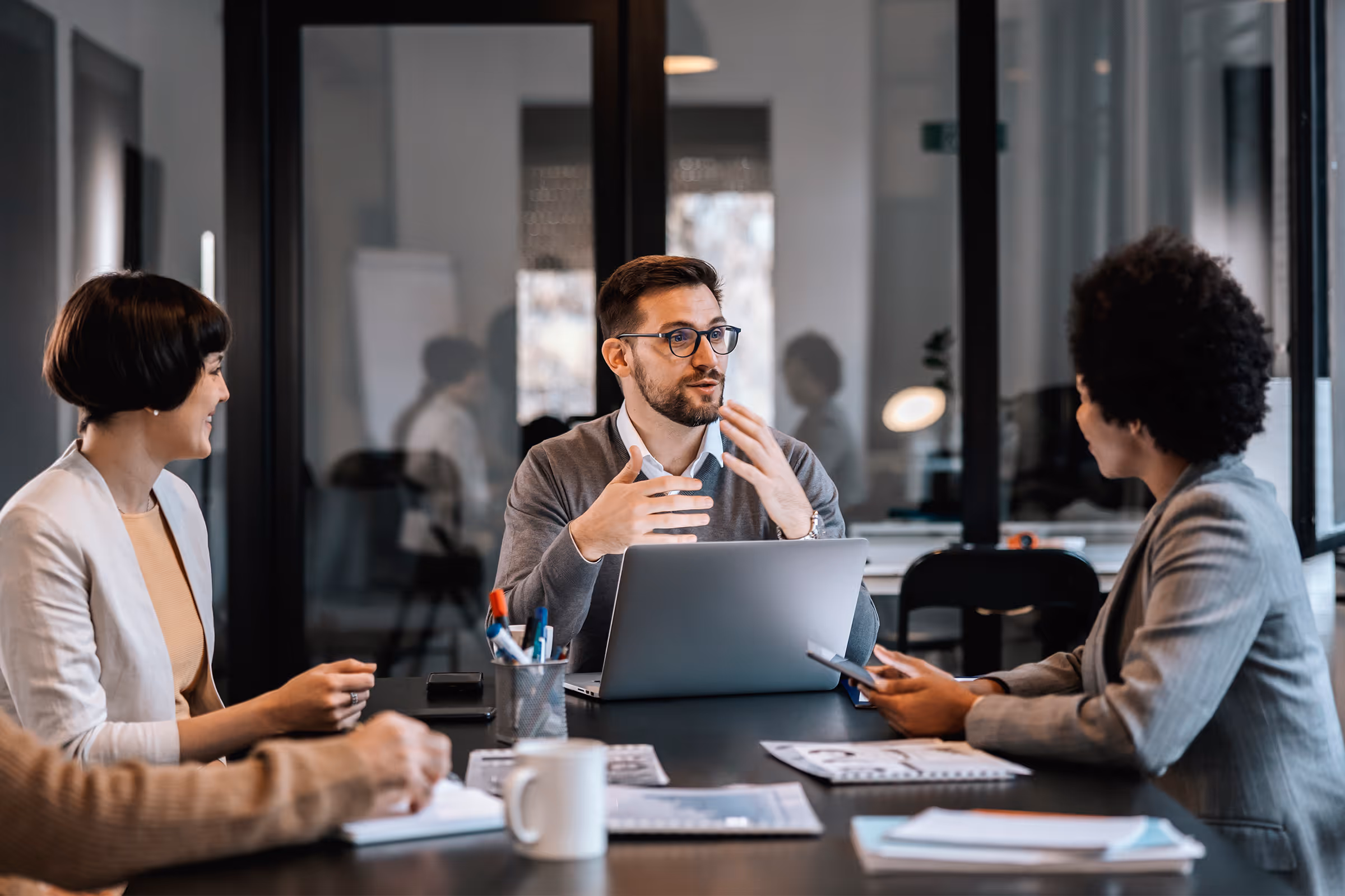 Three professionals having a discussion around a table in a modern office with a laptop and documents.