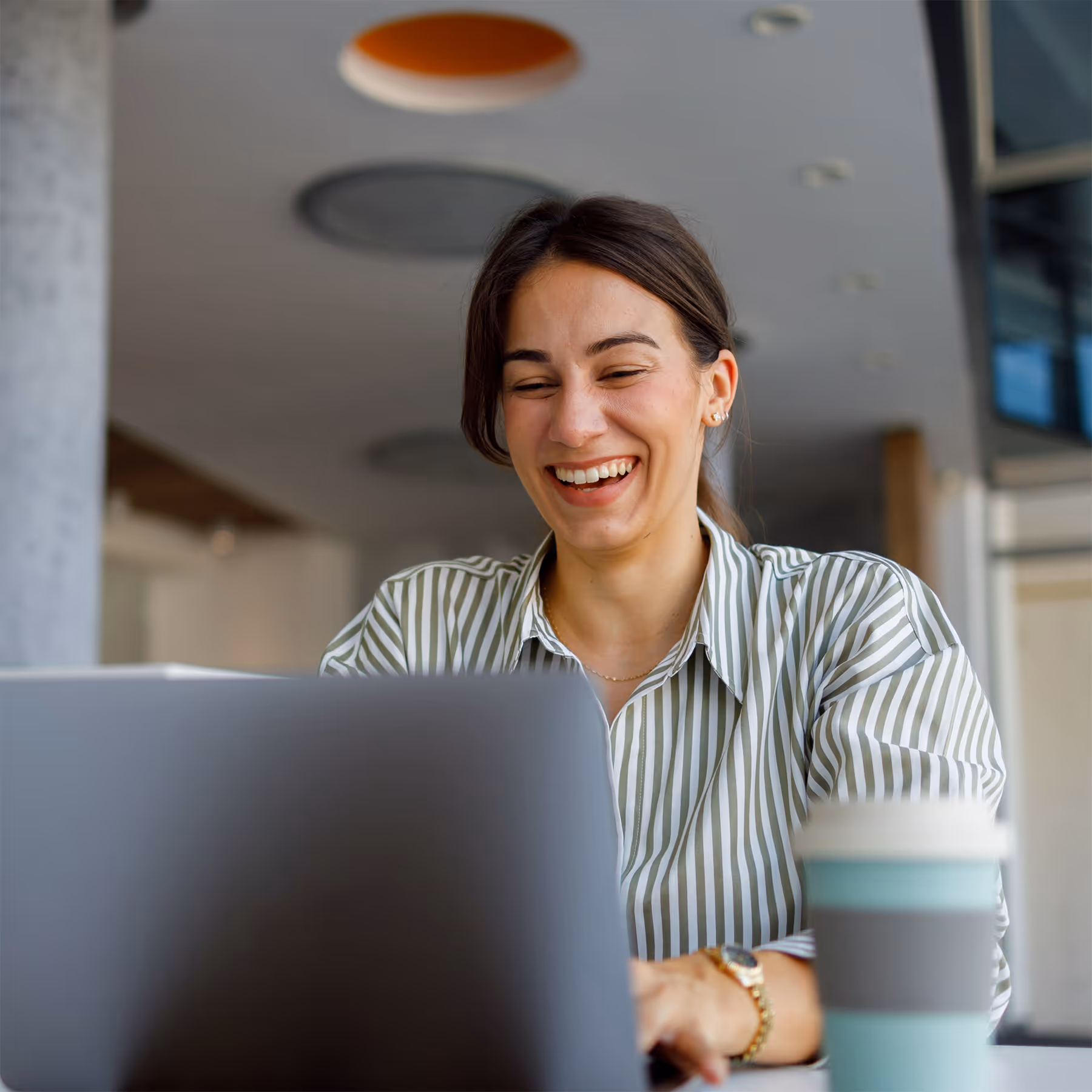 Smiling woman in a striped shirt working on a laptop with a reusable coffee cup nearby.